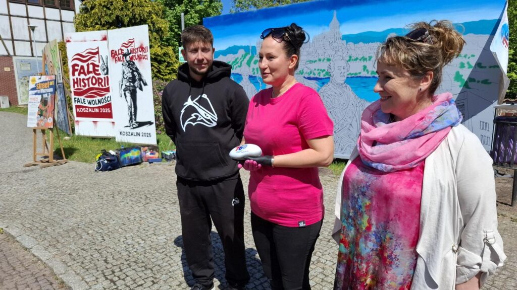 Trzy osoby stoją przed powstającym muralem podczas wydarzenia w muzeum. Kobieta w różowej koszulce przemawia do uczestników, obok stoją organizatorzy i banery promujące wydarzenie. (kliknięcie powiększy zdjęcie).