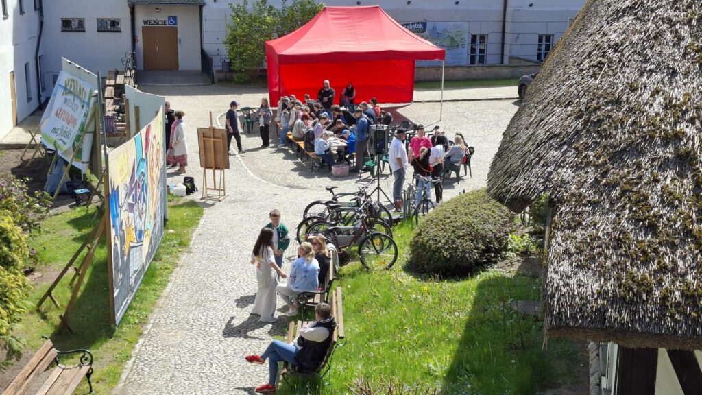 Widok z góry na dziedziniec muzeum podczas wydarzenia plenerowego. Uczestnicy siedzą pod czerwonym namiotem, a obok ustawiono plansze i duże graffiti. (kliknięcie powiększy zdjęcie).