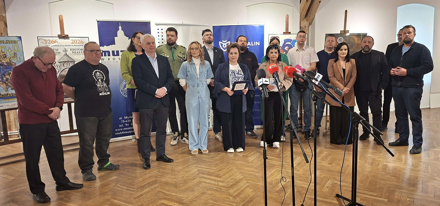 Goście konferencji prasowej pozują do wspólnego zdjęcia, stoją przed mikrofonami, na tle oprawionych w ramy plakatów.