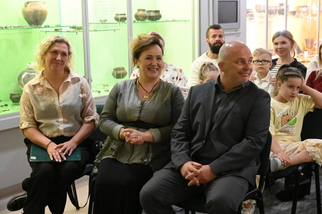 Uczestnicy wydarzenia siedzą na krzesłach w sali muzealnej. W tle widoczne są gabloty z eksponatami oraz pozostali goście konkursu (kliknięcie powiększy zdjęcie).