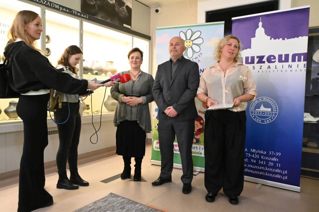obieta trzyma mikrofon podczas przemówienia otwierającego wydarzenie konkursowe w muzeum. Obok stoją organizatorzy na tle baneru muzeum (kliknięcie powiększy zdjęcie).