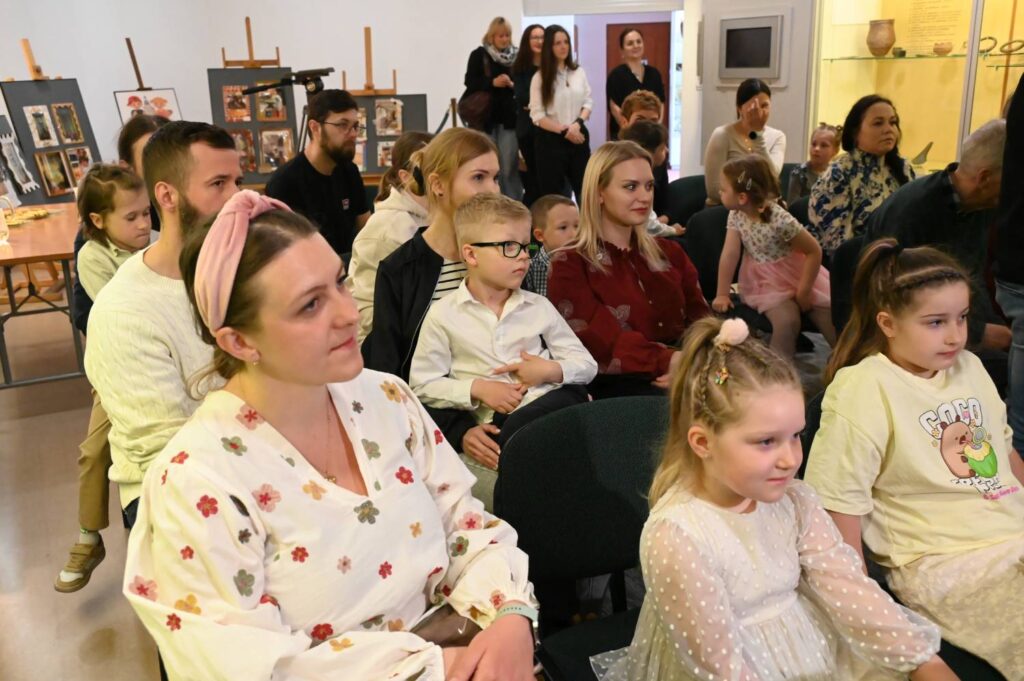 Publiczność, w tym dzieci i dorośli, uczestniczy w wydarzeniu konkursowym w sali muzealnej. W tle widoczne są prace prezentowane na sztalugach (kliknięcie powiększy zdjęcie).