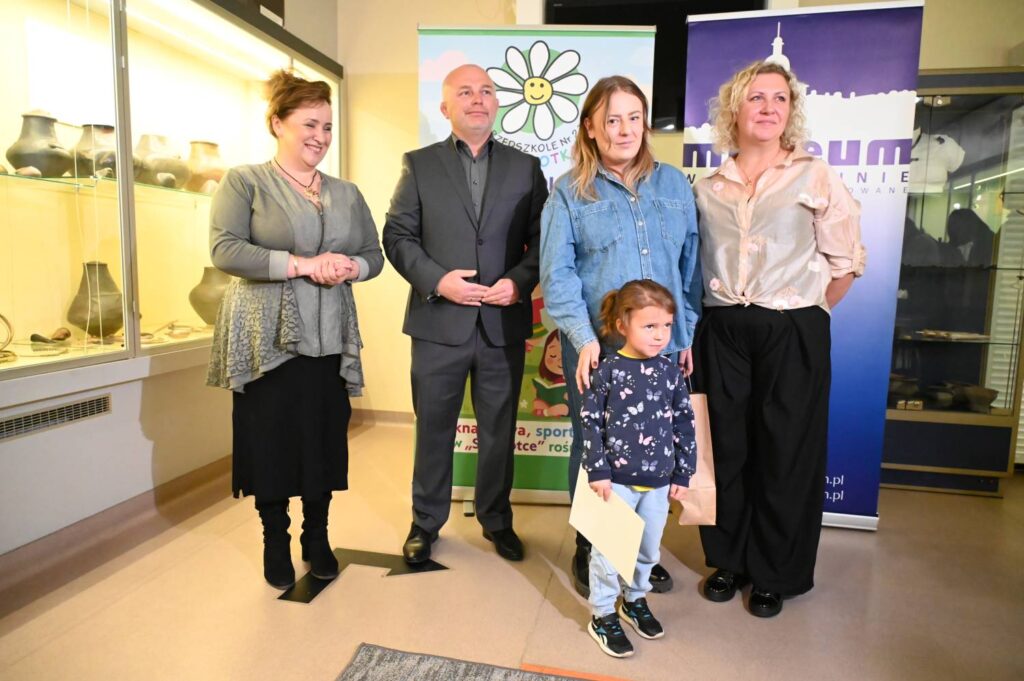 Dziewczynka – laureatka konkursu – pozuje do pamiątkowego zdjęcia z organizatorami wydarzenia w muzeum (kliknięcie powiększy zdjęcie).
