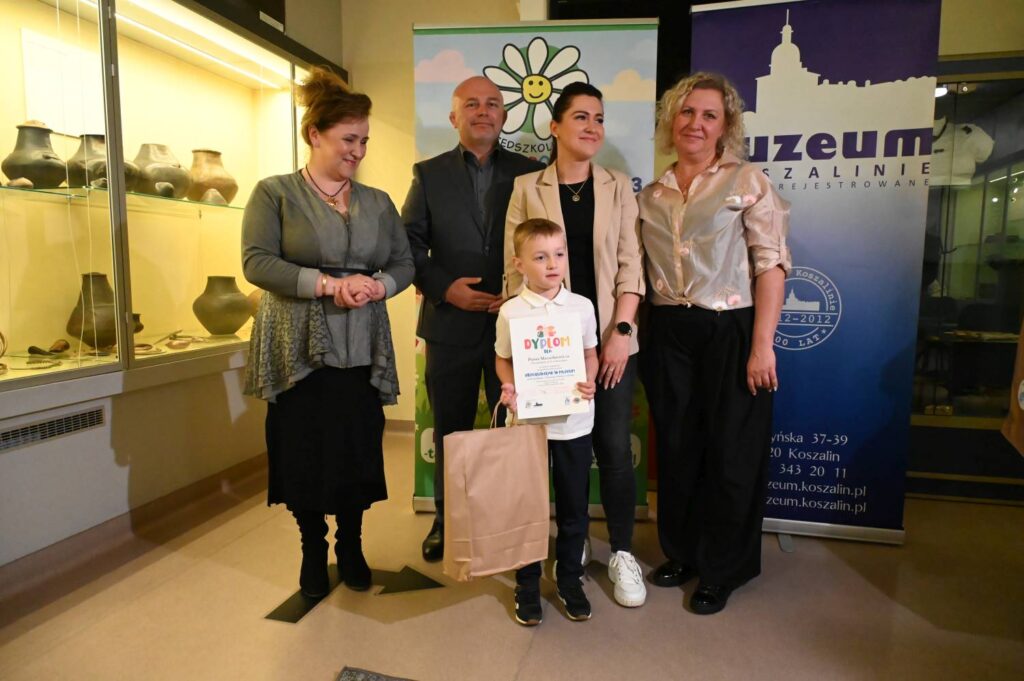 Chłopiec prezentuje dyplom i nagrodę podczas pamiątkowego zdjęcia z organizatorami konkursu (kliknięcie powiększy zdjęcie).