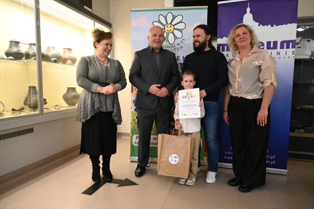 Dziewczynka prezentuje dyplom i nagrodę podczas pamiątkowego zdjęcia z organizatorami konkursu oraz opiekunem (kliknięcie powiększy zdjęcie).