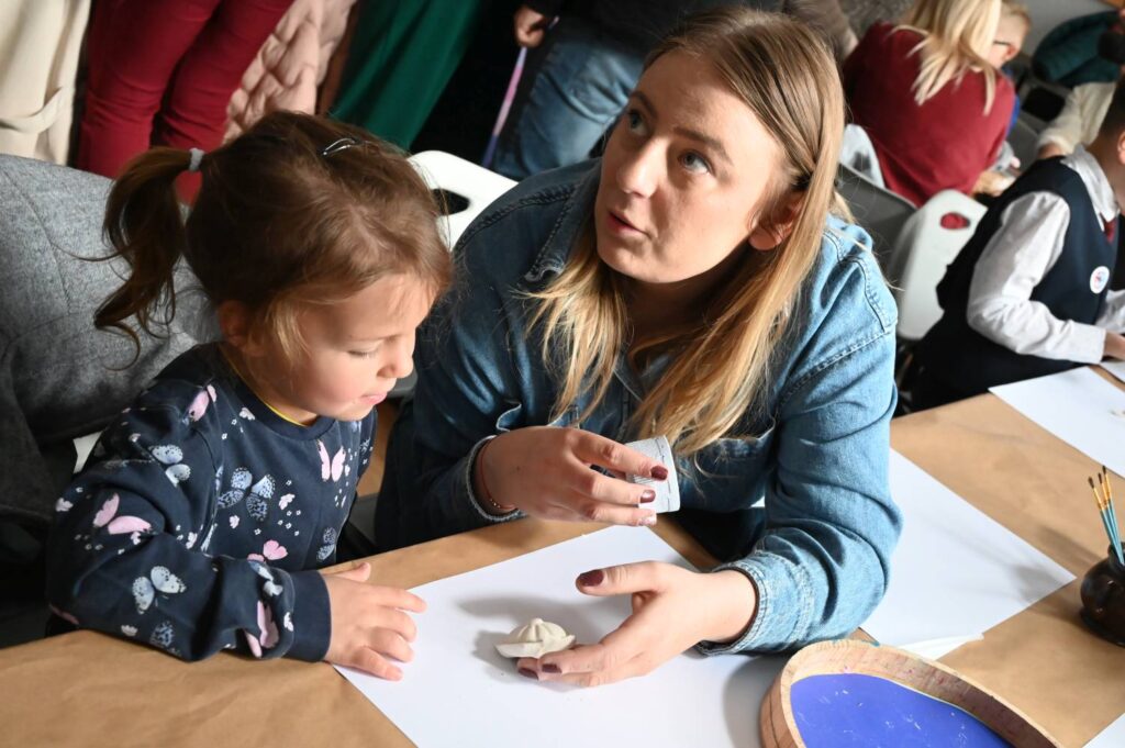 Kobieta rozmawia z dzieckiem podczas warsztatów artystycznych przy stole z materiałami plastycznymi (kliknięcie powiększy zdjęcie).