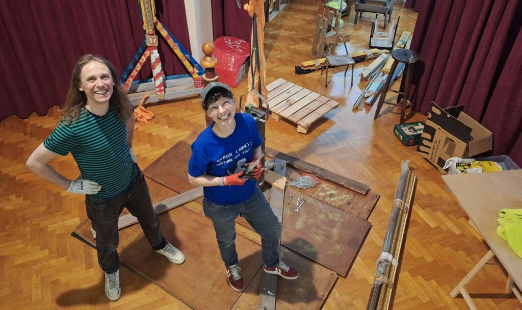 Dwie uśmiechnięte osoby stoją w sali podczas montażu scenografii lub instalacji artystycznej, wokół leżą drewniane elementy, narzędzia i materiały do budowy, w tle widoczne są bordowe kotary (kliknięcie powiększy zdjęcie).