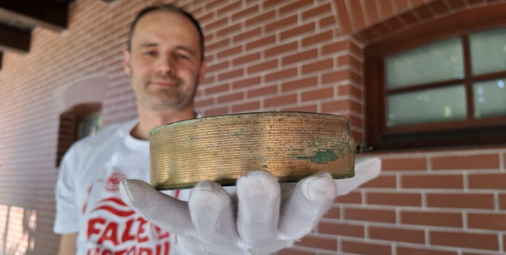 Archeolog prezentuje brązowy naszyjnik, trzymany na dłoni w białej rękawiczce, w tle stylizowana na czerwoną cegłę ściana budynku (kliknięcie powiększy zdjęcie).