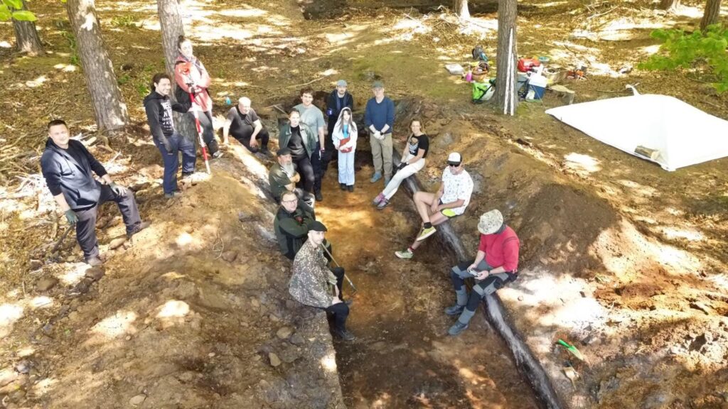 Grupa archeologów i uczestników badań zgromadzona wokół wykopu podczas omawiania przebiegu prac terenowych (kliknięcie powiększy zdjęcie).