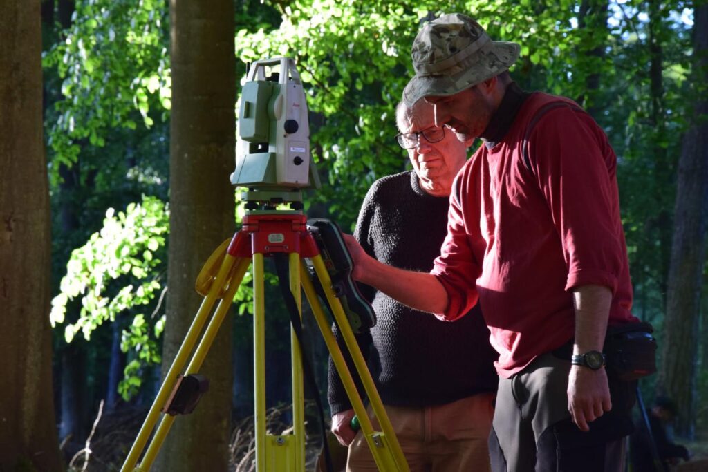 Dwie osoby stoją przy urządzeniu geodezyjnym i wykonują pomiary stanowiska archeologicznego przy użyciu tachimetru (kliknięcie powiększy zdjęcie).