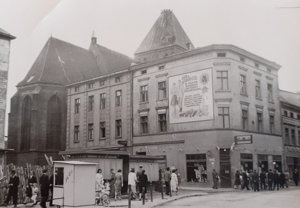 Historyczna zabudowa miejska z kamienicą przy skrzyżowaniu ulic, widoczni przechodnie oraz przystanek. (kliknięcie powiększy zdjęcie).
