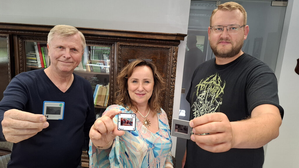 Trzy osoby stoją w biurze, obok siebie i patrzą w kierunku obiektywu, każda z nich trzyma w dłoni jeden slajd.