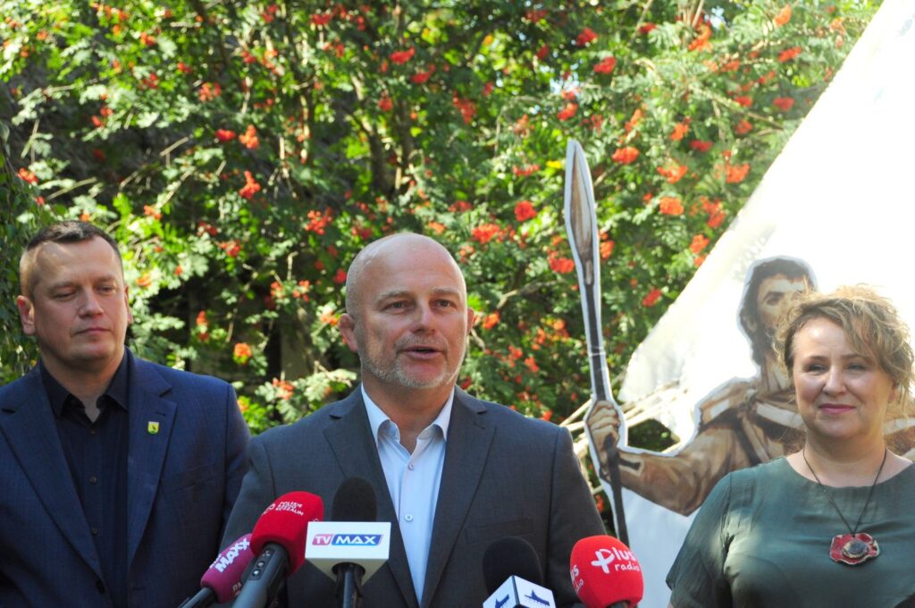 Trzy osoby uczestniczą w konferencji prasowej; przed nimi ustawiono mikrofony z logotypami stacji telewizyjnych, a w tle widoczny jest namiot i element scenografii wydarzenia (kliknięcie powiększy zdjęcie).