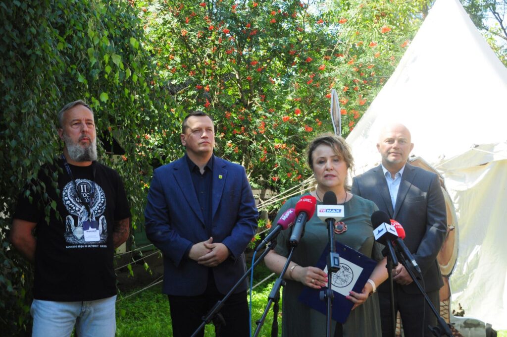 Kobieta przemawia podczas konferencji prasowej; przed nią znajdują się mikrofony kilku redakcji, a za nią stoją pozostali uczestnicy spotkania (kliknięcie powiększy zdjęcie).