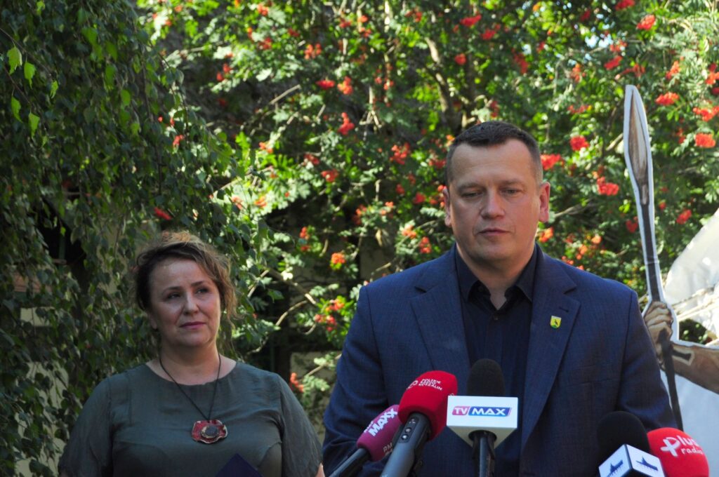Kobieta i mężczyzna uczestniczą w konferencji prasowej; mężczyzna przemawia do mikrofonów ustawionych przed nimi, a w tle widoczny jest element scenografii wydarzenia (kliknięcie powiększy zdjęcie).