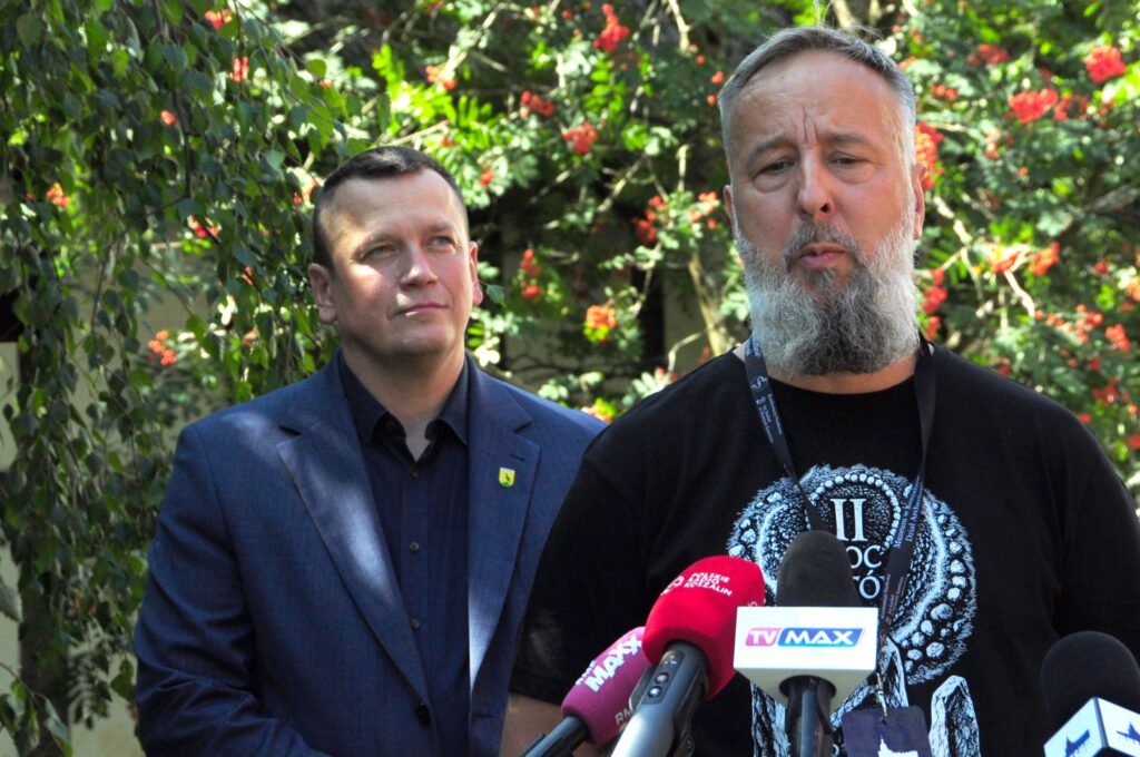 Dwóch mężczyzn uczestniczy w konferencji prasowej; jeden z nich stoi przy mikrofonach i przemawia, a w tle widoczna jest porośnięta zielenią ściana (kliknięcie powiększy zdjęcie).
