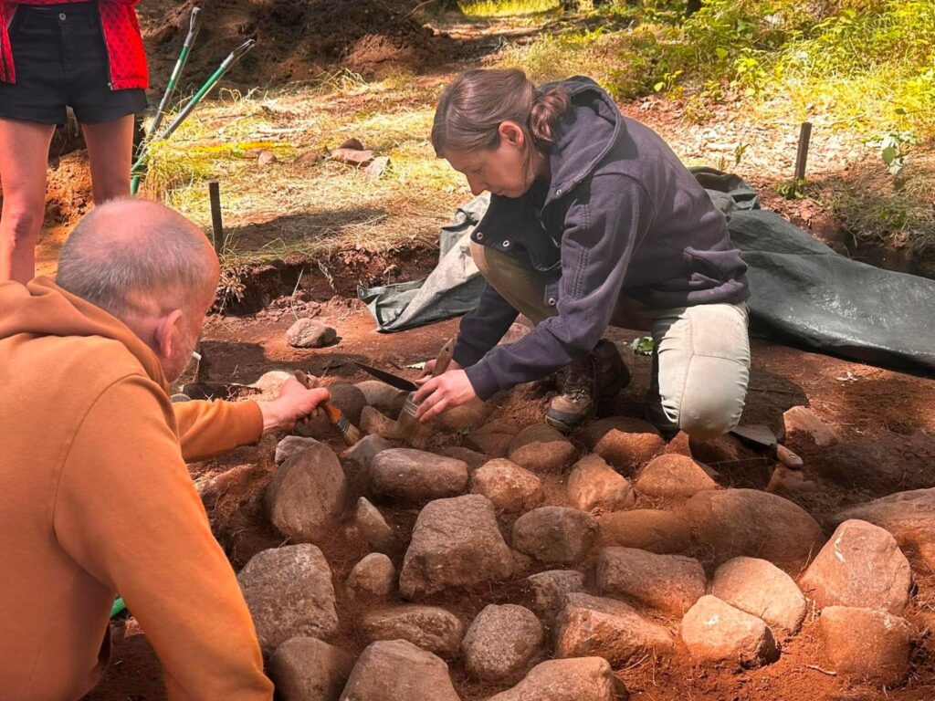Osoba będąca w wykopie  oczyszcza odsłoniętą kamienną konstrukcję, usuwając ziemię spomiędzy otoczaków. Obok niej druga osoba przygląda się postępom prac i pomaga w dokumentowaniu odkrycia (kliknięcie powiększy zdjęcie).