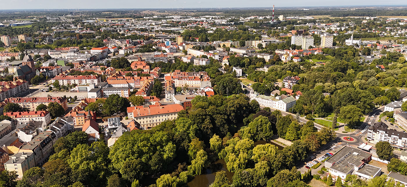 Panoramiczne ujęcie Koszalina z lotu ptaka; w centrum miasta widoczna jest zabudowa z czerwonymi dachami, rozległy park z licznymi drzewami i stawem oraz otaczające miasto osiedla i tereny zielone; w oddali widoczne są pola, lasy, elektrownia wiatrowa i komin przemysłowy (kliknięcie powiększy zdjęcie).