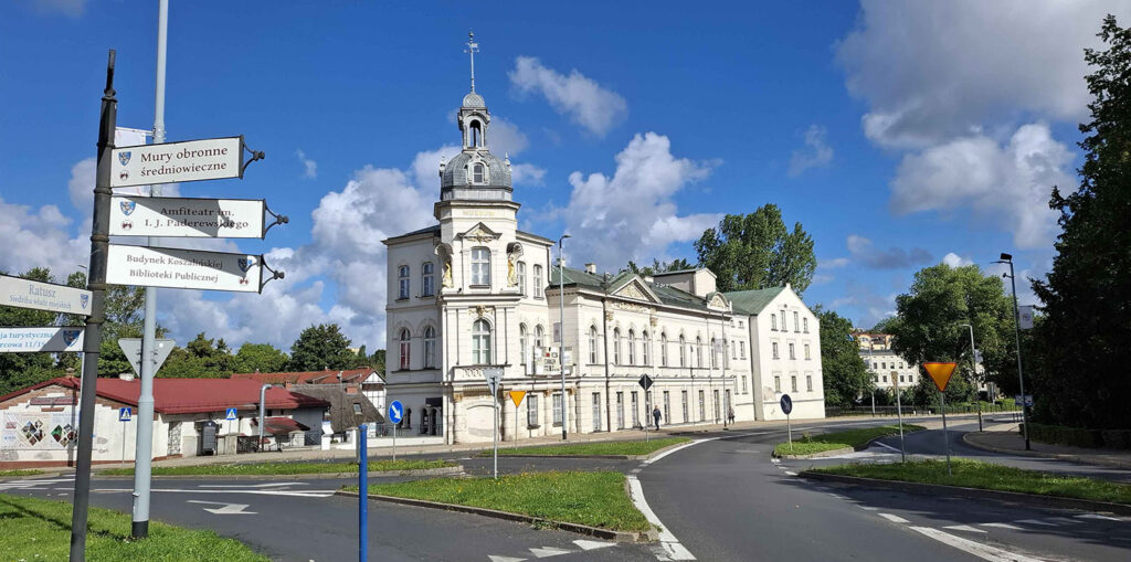 Budynek obecnego Muzeum w Koszalinie widziany od strony ulicy; przed gmachem znajduje się rondo z drogowskazami wskazującymi m.in. mury obronne, amfiteatr i Bibliotekę Publiczną; budynek ma charakterystyczną wieżę z kopułą, a w tle widoczne są drzewa i zabudowa miasta (kliknięcie powiększy zdjęcie).