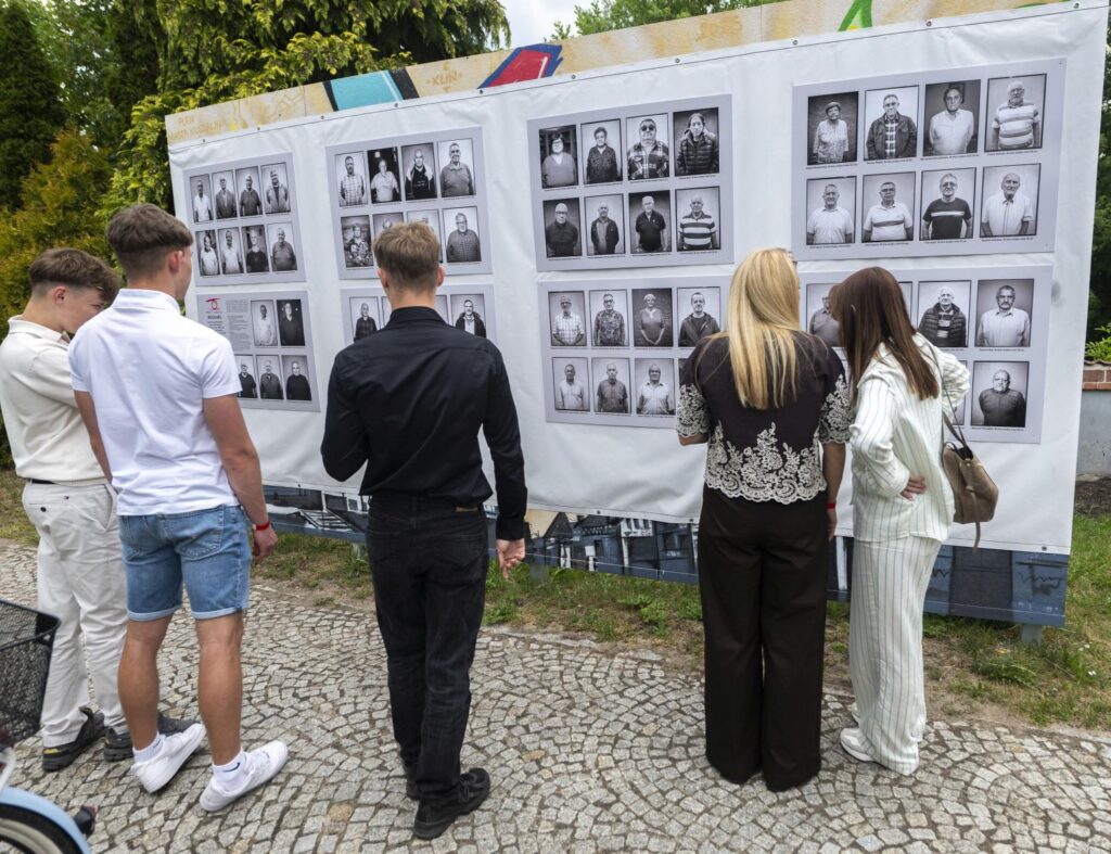 Kilka osób stoi tyłem do obiektywu i ogląda plenerową wystawę czarno-białych portretów przedstawiających różne osoby (kliknięcie powiększy zdjęcie).