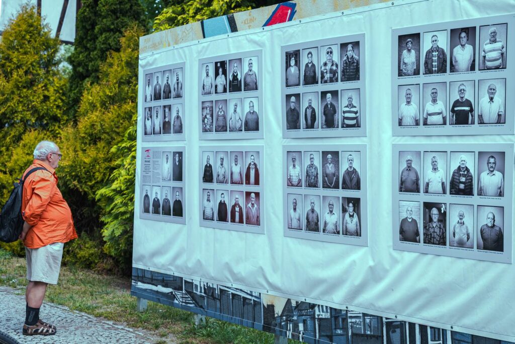 Mężczyzna ogląda tablice ekspozycyjne z czarno-białymi fotografiami portretowymi; w tle drzewa i krzewy (kliknięcie powiększy zdjęcie).