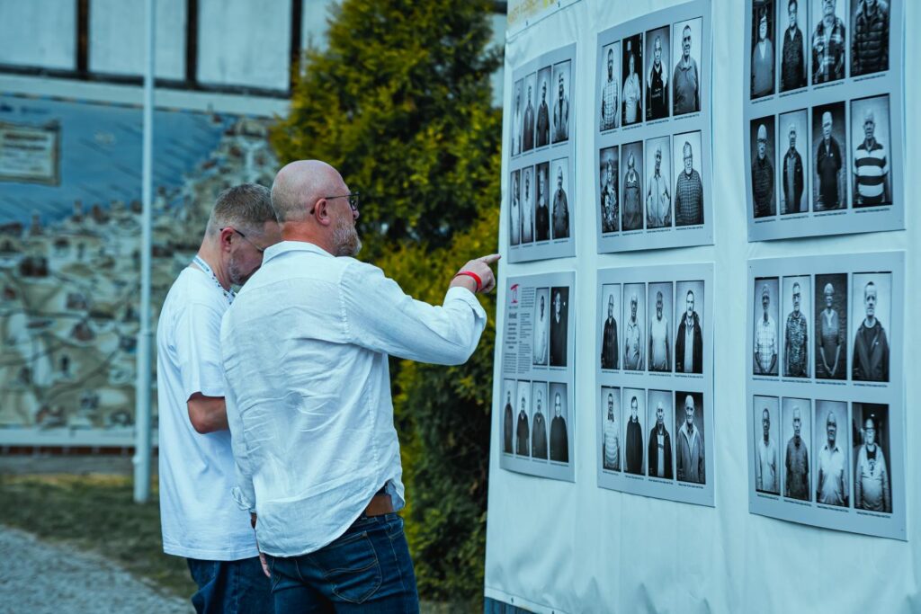 Dwóch mężczyzn przygląda się z bliska czarno-białym portretowym fotografiom prezentowanym na plenerowej wystawie; w tle fragment ściany budynku stylizowanej na wzór konstrukcji ryglowej (kliknięcie powiększy zdjęcie).