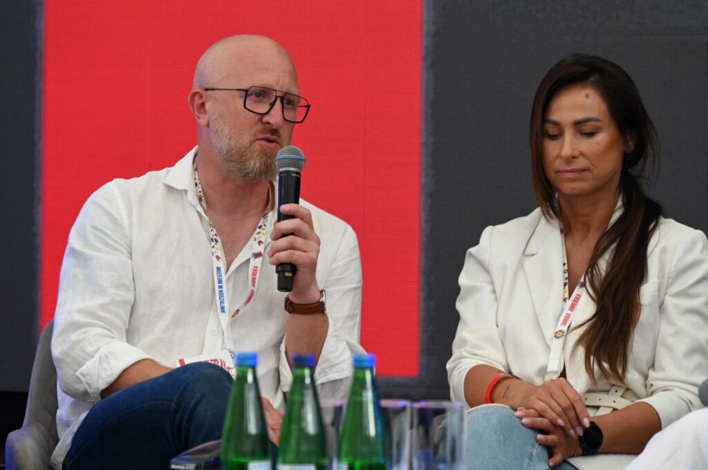 Dwoje uczestników panelu dyskusyjnego rozmawia podczas festiwalu „Fale Historii”. Jeden z gości zabiera głos do mikrofonu, a druga uczestniczka przysłuchuje się jego wypowiedzi (kliknięcie powiększy zdjęcie)